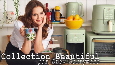 Drew Barrymore sourit en tenant une tasse colorée dans une cuisine au style moderne. Elle est entourée d'appareils électroménagers vert sauge de sa "Collection Beautiful", incluant une friteuse à air, une cafetière, une bouilloire et un grille-pain four. Le décor est lumineux et chaleureux, avec des touches de plantes et d'aliments frais, créant une ambiance conviviale.